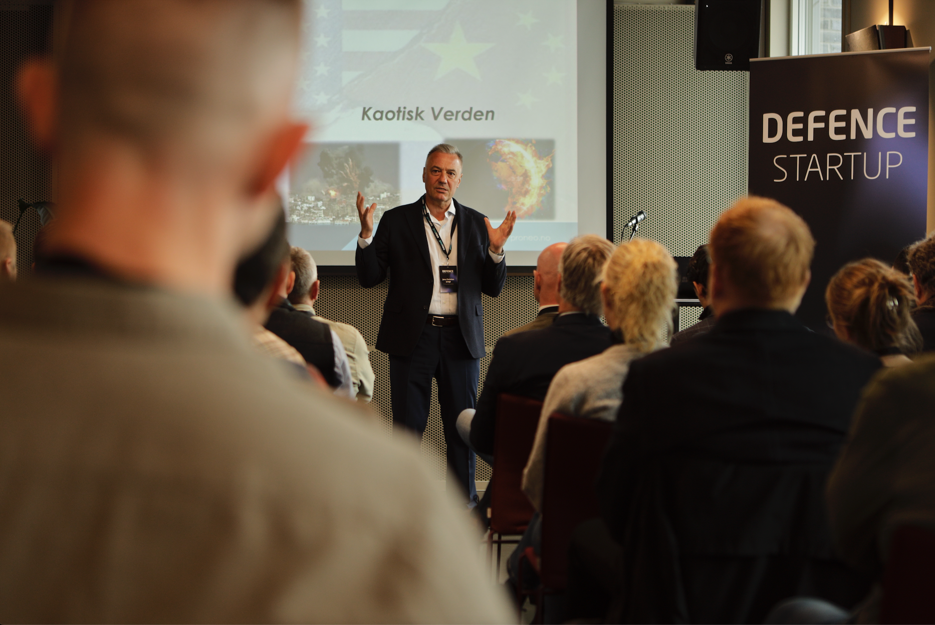 Male speaker in a dark suit presenting to a seated audience at a conference with a projection screen behind him showing 'Kaotisk Verden' and a 'Defence Startup' banner nearby