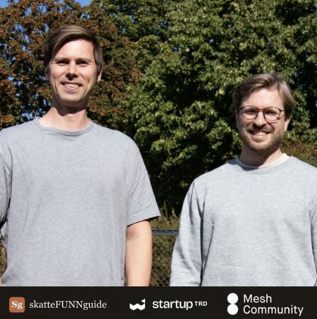 Two men in gray shirts stand outdoors, smiling at the camera with trees in the background.